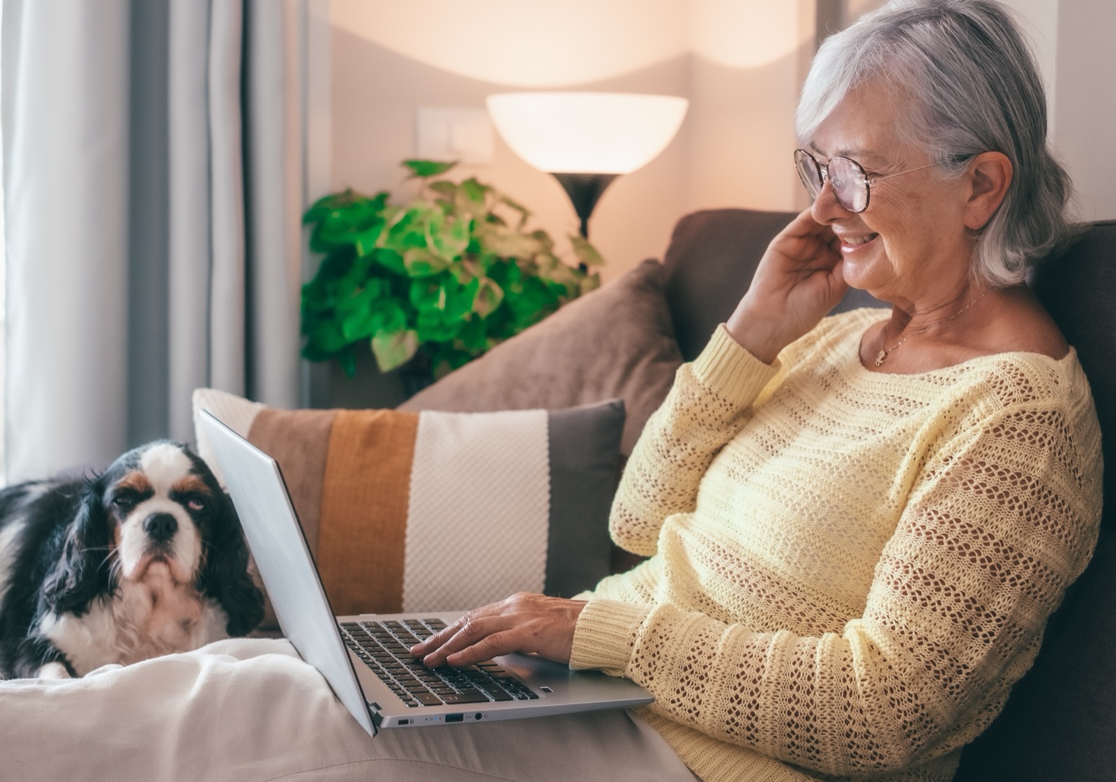 An elderly woman smiling at her laptop with her dog beside her